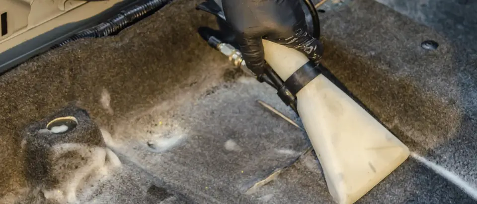 A hand in a black glove pressing the clear nozzle of an extraction vacuum onto wet grey carpet with patches of foam