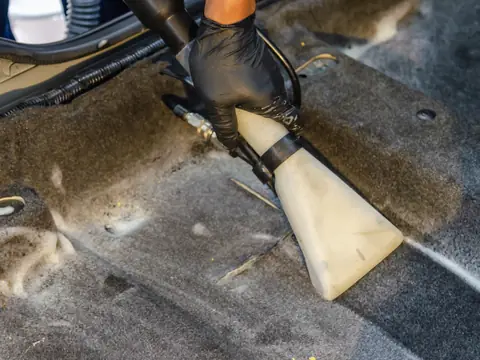 A hand in a black glove pressing the clear nozzle of an extraction vacuum onto wet grey carpet with patches of foam