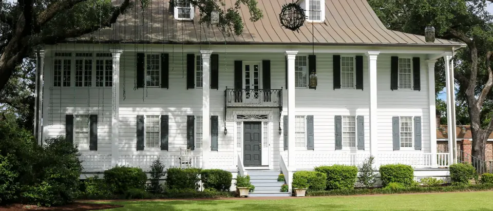 A two-storey white frame house with dark shutters and a full-width porch on square columns, seen across a lawn under old oak trees